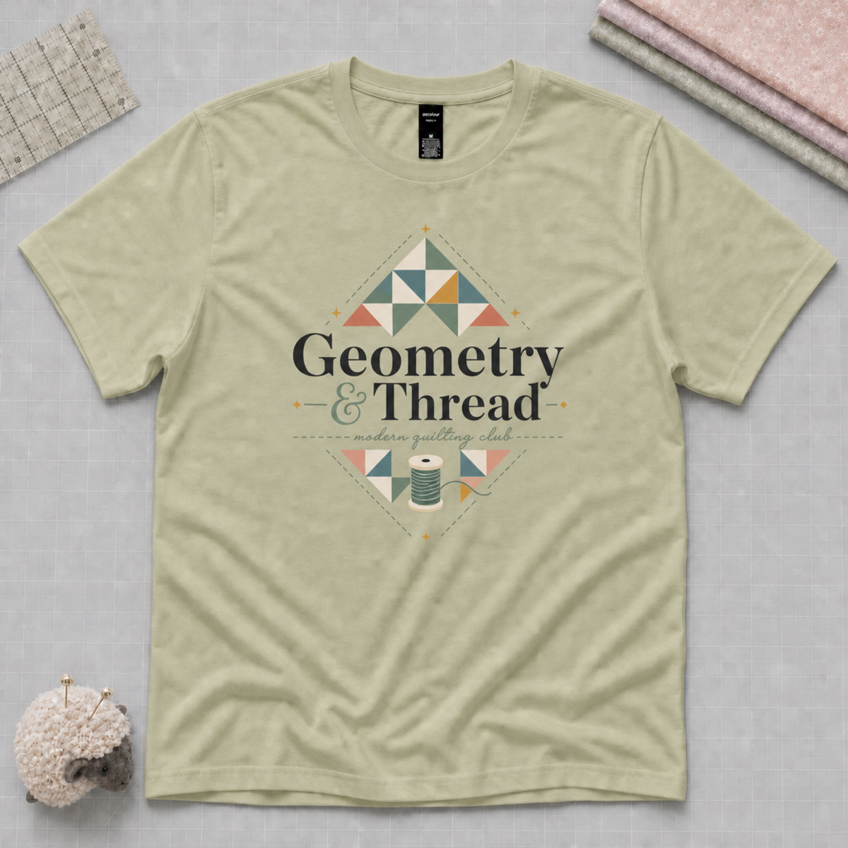 A pistachio green short-sleeve t-shirt laying flat on a pale grey cutting mat withe quilting tools in the corners. The t-shirt features a quilt-themed graphic with the words 'Geometry & Thread' surrounded by patchwork triangles.