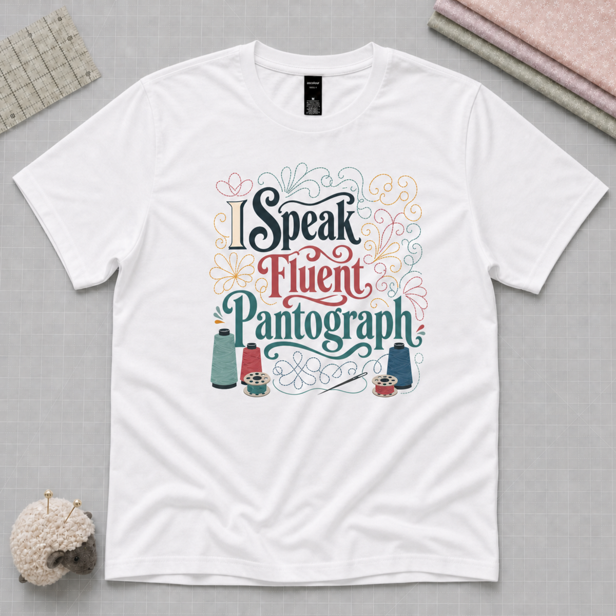 A white short-sleeve T-shirt featuring the phrase 'I Speak Fluent Pantograph' in decorative multicolour lettering with sewing-themed illustrations. Styled flat-lay props include a quilting ruler, folded fabric stack, and a small pincushion.