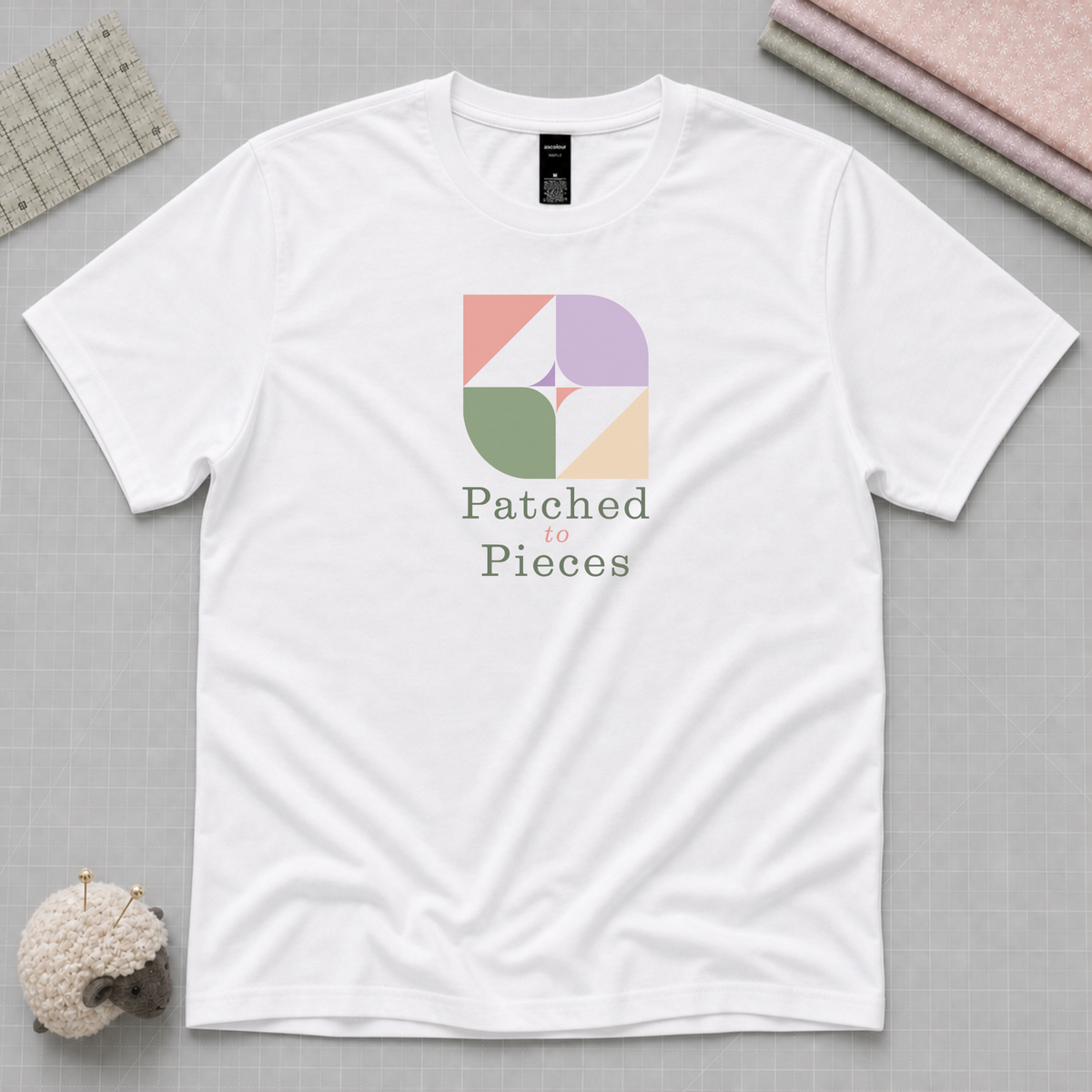 A white short-sleeve crewnect t-shirt with the 'Patched to Pieces' Logo laying flat on a pale grey cutting mat with sewing accessories in the corners.