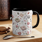 A ceramic mug with a white exterior decorated with a vintage-style pattern of flowers, hearts, and fabric, paired with a black interior and handle. It is styled on a wooden surface next to a spoon and a macaron, with a blurred background.