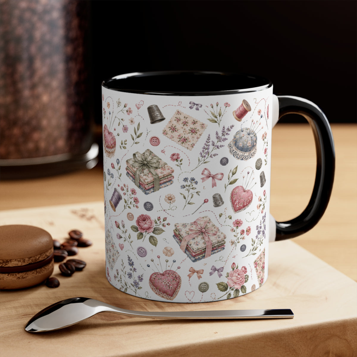 A ceramic mug with a white exterior decorated with a vintage-style pattern of flowers, hearts, and fabric, paired with a black interior and handle. It is styled on a wooden surface next to a spoon and a macaron, with a blurred background.