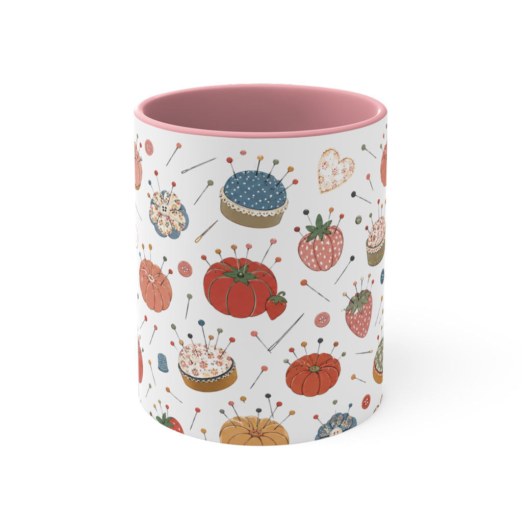 A white ceramic mug with a pink interior, decorated with a vintage-style pin cushion pattern featuring red pumpkins, strawberries, and pincushions on a white background.