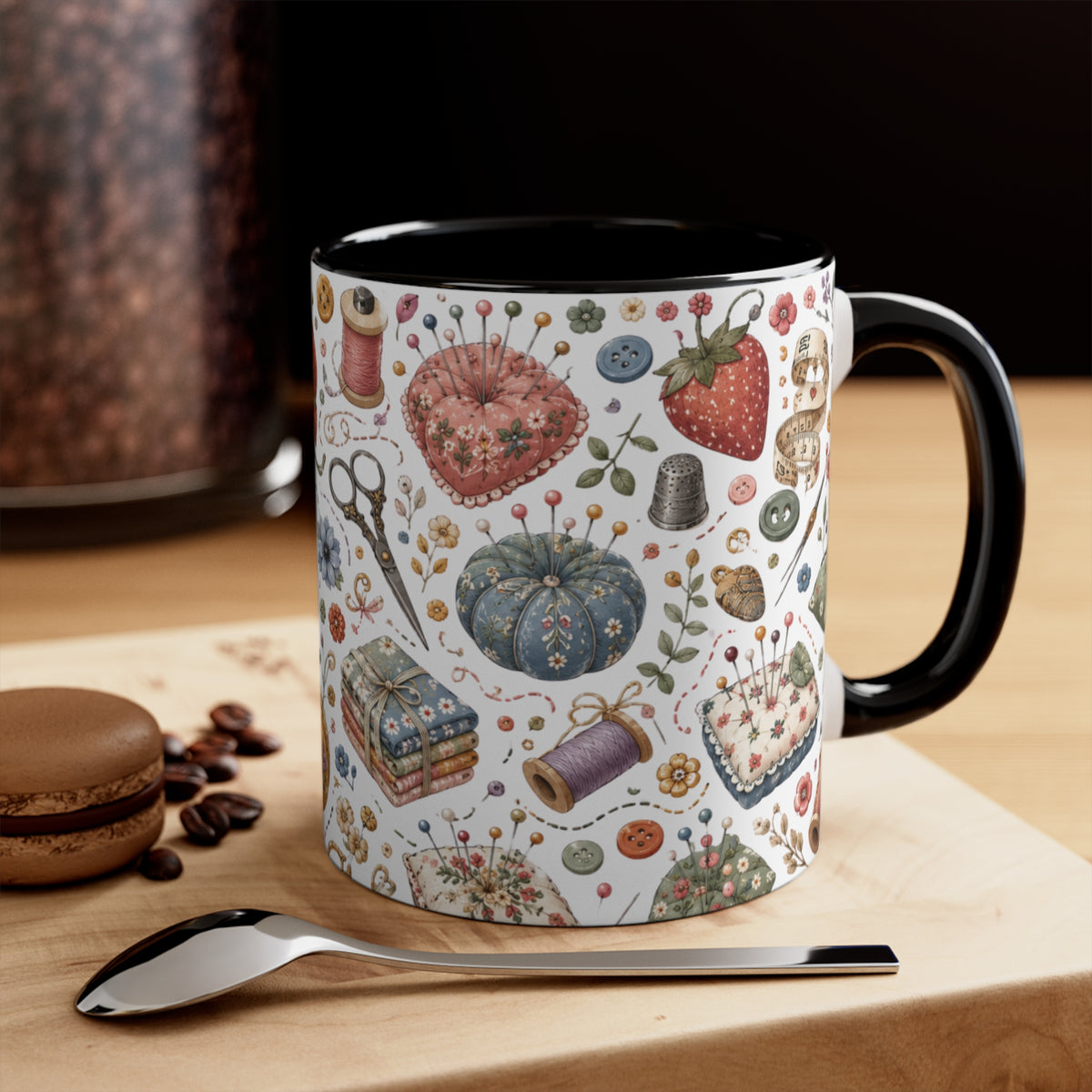 A ceramic mug with a white base decorated with vintage-style sewing and craft illustrations, including pincushions, scissors and thread spools, with a black handle and interior. It is shown on a wooden surface with a spoon in front.