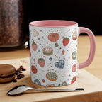 A white ceramic mug with a pink handle and interior, decorated with a whimsical pattern of strawberries, pumpkins, pins, and sewing notions, displayed on a wooden board with a spoon and a macaron in the background.