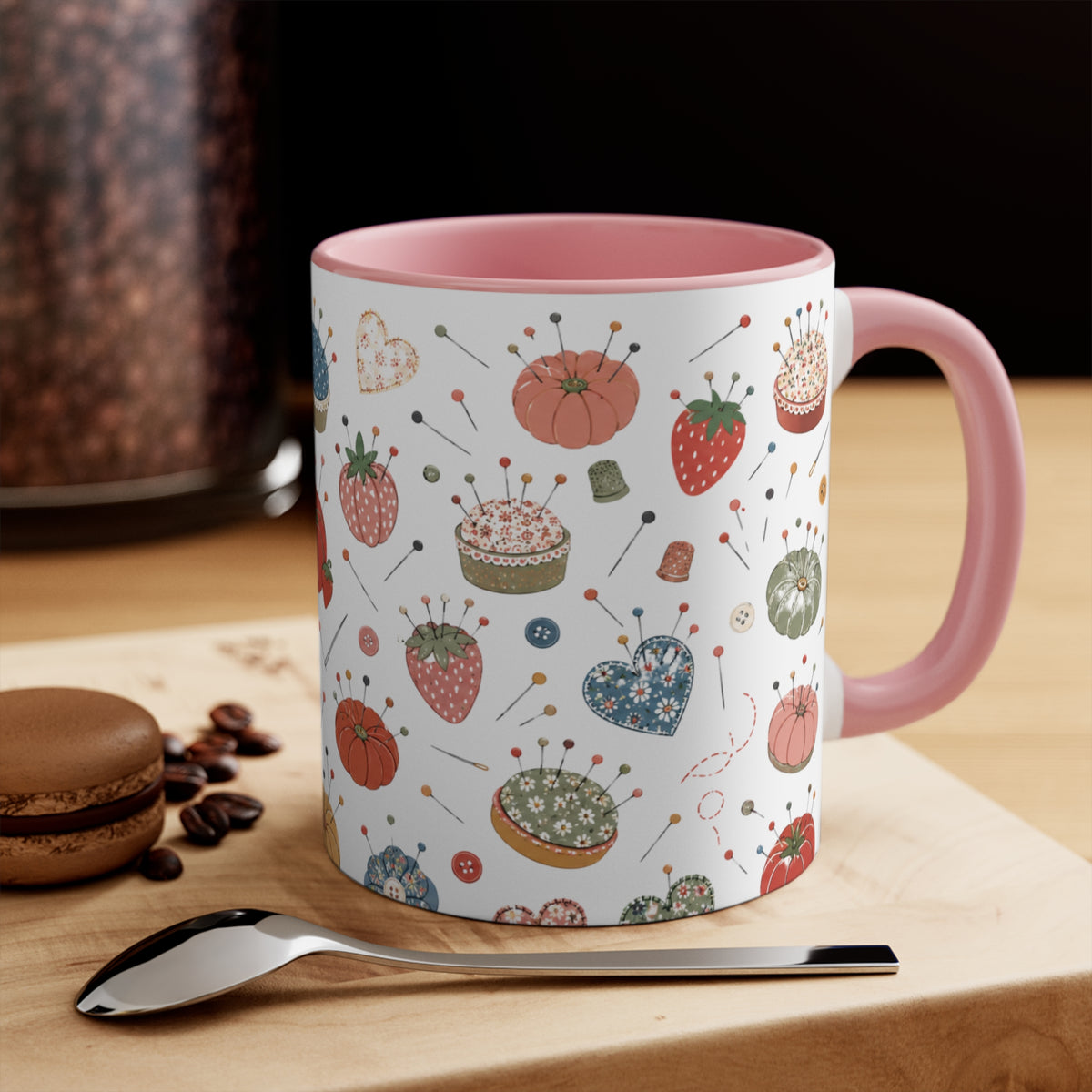 A white ceramic mug with a pink handle and interior, decorated with a whimsical pattern of strawberries, pumpkins, pins, and sewing notions, displayed on a wooden board with a spoon and a macaron in the background.