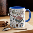 A white ceramic mug with a blue handle and blue interior, decorated with quilting-themed illustrations including fabric stacks, a coffee cup, thread spools, and a quilt pattern. The mug sits on a wooden surface with a spoon in front and a blurred coffee jar in the background.