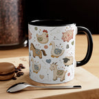 A white ceramic mug with a black handle and interior, decorated with a whimsical pincushion animals including chickens, pigs, ducks, horses, sheep, and goats. The mug is styled on a wooden board with a spoon, coffee beans, and a macaron in the background.