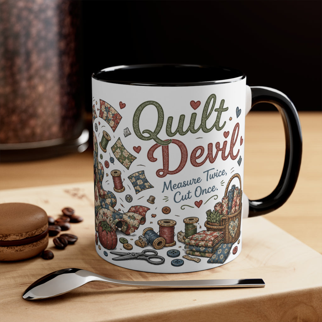 A white ceramic mug with a black handle and interior, decorated with a vintage-style quilt design featuring the words 'Quilt Devil' and 'Measure Twice, Cut Once.' The mug is styled on a wooden surface with a spoon, coffee beans, and a macaron in the background.