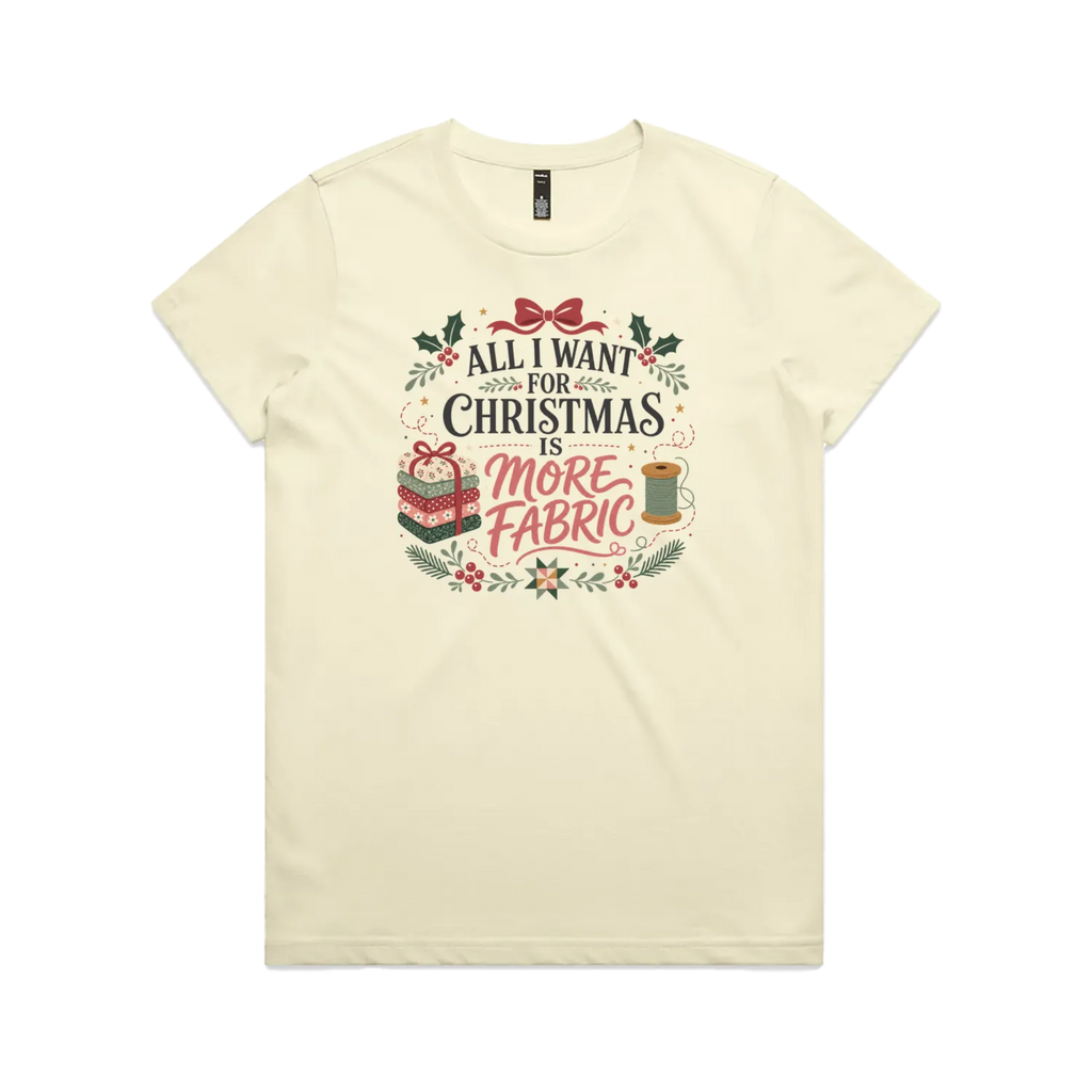 A light butter yellow short-sleeve crewneck t-shirt with a festive Christmas graphic on the front. The graphic features the text 'ALL I WANT FOR CHRISTMAS IS MORE FABRIC' in a mix of black and red fonts, surrounded by illustrations of a ribbon-tied fabric stack, a thread spool, and holly leaves.