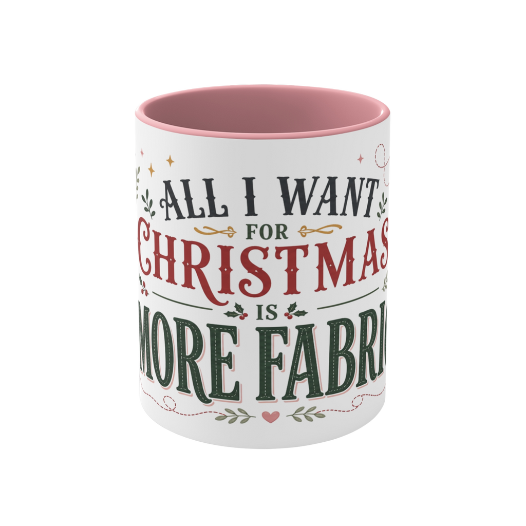 The front of a white ceramic mug with a pink interior and rim, showing the words "All I Want for Christmas is More Fabric'. The text is styled in dark green and red fonts with small holly and heart accents.