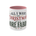 The front of a white ceramic mug with a pink interior and rim, showing the words "All I Want for Christmas is More Fabric'. The text is styled in dark green and red fonts with small holly and heart accents.