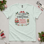 A pale seafoam green t-shirt laying flat on a grey cutting mat with quilting equipment and holly leaves in the corners. The graphic reads 'All I Want For Christmas is More Fabric, with a stack of fabric, a thread reel and leaves around the writing.