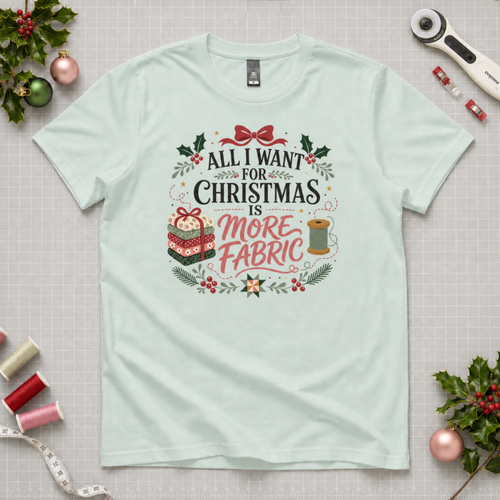 A pale seafoam green t-shirt laying flat on a grey cutting mat with quilting equipment and holly leaves in the corners. The graphic reads 'All I Want For Christmas is More Fabric, with a stack of fabric, a thread reel and leaves around the writing.