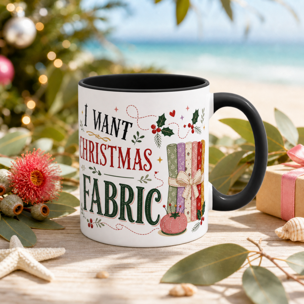 A white ceramic mug with a black handle and interior, printed with the phrase 'All I Want for Christmas is More Fabric' in black, red and green lettering alongside a colorful quilt illustration. The mug is styled on a wooden surface with eucalyptus leaves, a starfish, a pink flower, and a blurred beach and Christmas tree background.