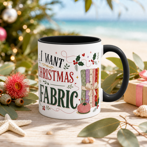 A white ceramic mug with a black handle and interior, printed with the phrase 'All I Want for Christmas is More Fabric' in black, red and green lettering alongside a colorful quilt illustration. The mug is styled on a wooden surface with eucalyptus leaves, a starfish, a pink flower, and a blurred beach and Christmas tree background.