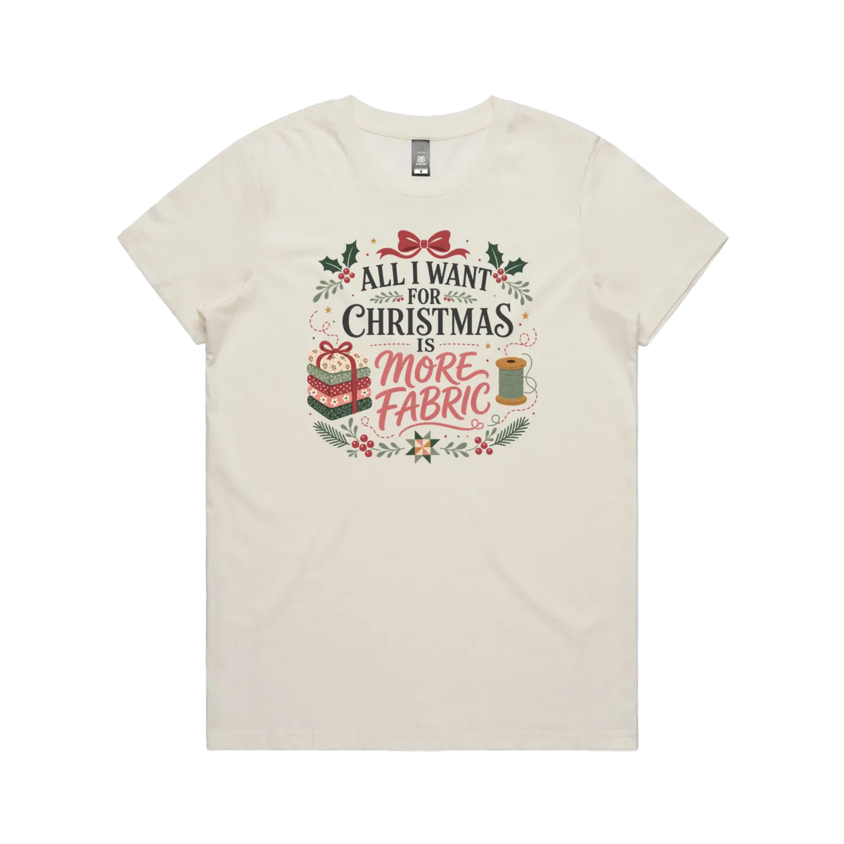 An off-white short-sleeve crewneck t-shirt with a festive Christmas graphic on the front. The graphic features the text 'ALL I WANT FOR CHRISTMAS IS MORE FABRIC' in a mix of black and red fonts, surrounded by illustrations of a ribbon-tied fabric stack, a thread spool, and holly leaves.