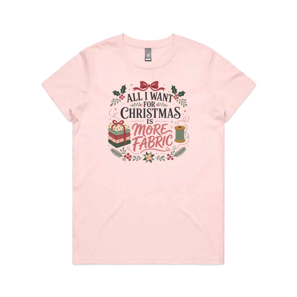 A light pink short-sleeve crewneck t-shirt with a festive Christmas graphic on the front. The graphic features the text 'ALL I WANT FOR CHRISTMAS IS MORE FABRIC' in a mix of black and red fonts, surrounded by illustrations of a ribbon-tied fabric stack, a thread spool, and holly leaves.