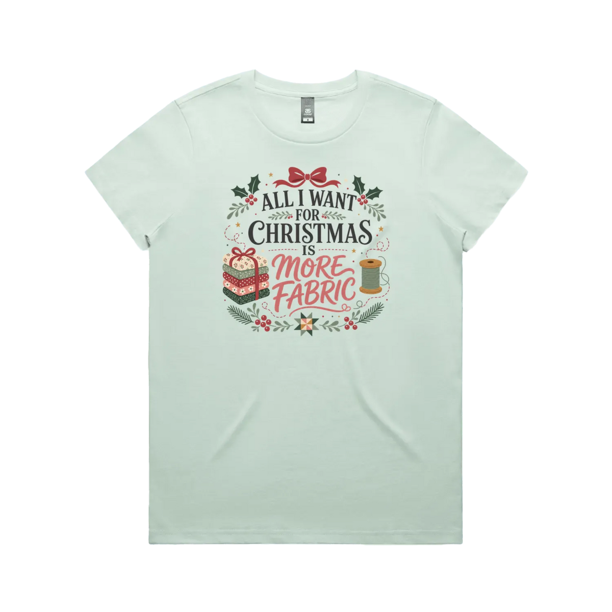 A light seafoam green short-sleeve crewneck t-shirt with a festive Christmas graphic on the front. The graphic features the text 'ALL I WANT FOR CHRISTMAS IS MORE FABRIC' in a mix of black and red fonts, surrounded by illustrations of a ribbon-tied fabric stack, a thread spool, and holly leaves.