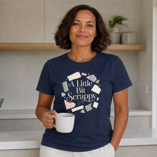 A woman with short dark hair stands in a kitchen setting, wearing a navy blue graphic t-shirt that reads 'A Little Bit Scrappy' with a colorful fabric scrap themed design. She is holding a white ceramic mug.