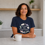 A woman with short dark hair sits at a light gray table in a bright kitchen, wearing a navy blue graphic T-shirt that reads 'Little Bit Scrappy' with colorful fabric swatch illustrations. A white mug sits in front of her.