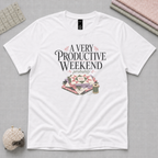 A white short-sleeve graphic t-shirt laying flat on a pale grey background with quilting supplies in the corners. The shirt reads 'A Very Productive Weekend, probably' above a stack of quilt blocks and a thread spool.