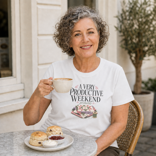 An older woman with short grey hair sits at an outdoor table wearing a white graphic t-shirt with the words 'A Very Productive Weekend, probably'. She is holding a cup of coffee and there is a plate of scones, jam and cream on the table in front of her.