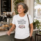 An older woman with short grey hair stands at a cafe counter. She is wearing a white graphic t-shirt with the words 'A Very Productive Weekend, probably' with dark pants. There are cakes on the counter and a chalkboard on the wall in the background.