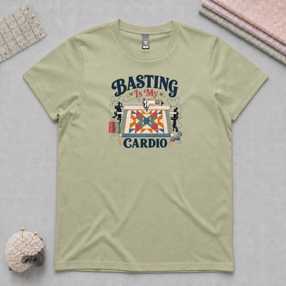 A pistachio green short-sleeve t-shirt laying on a pale grey cutting mat with quilting supplies in the corners. The graphic on the t-shirt reads 'Basting Is My Cardio' with an illustration of a longarm quilting machine, a patchwork quilt and thread.