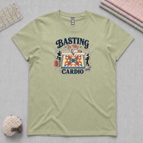 A pistachio green short-sleeve t-shirt laying on a pale grey cutting mat with quilting supplies in the corners. The graphic on the t-shirt reads 'Basting Is My Cardio' with an illustration of a longarm quilting machine, a patchwork quilt and thread.