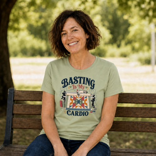 A woman with short brown hair sitting on a bench in a park wearing a pistachio green short-sleeve tshirt. The graphic on the t-shirt reads 'Basting Is My Cardio' with an illustration of a longarm quilting machine, a patchwork quilt and thread.