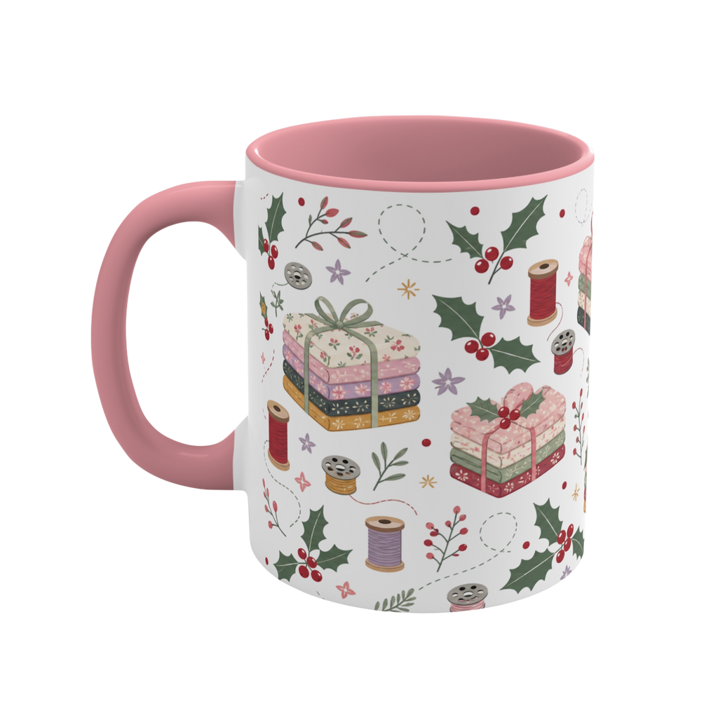 The left side of a white ceramic mug with a pink handle and interior, decorated with a repeating pattern of stacked fabrics, holly leaves, berries, and thread spools in soft pink, green, beige, and brown tones.