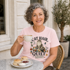 A smiling woman with curly gray hair sits at an outdoor table holding a mug, wearing a light pink graphic T-shirt that reads 'Cat Hair is Quilt Glitter' with an illustration of a cat and sewing motifs. A plate with scones and jam is on the table in front of her.