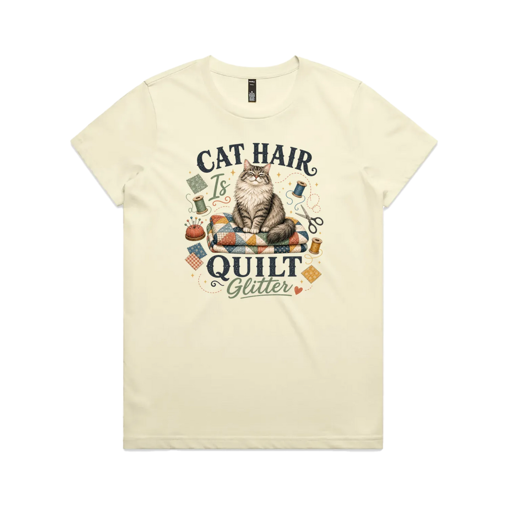 A pale butter yellow short-sleeve t-shirt with the words 'Cat Hair is Quilt Glitter' around an illustration of a cat sitting on a quilt surrounded by sewing accessories.