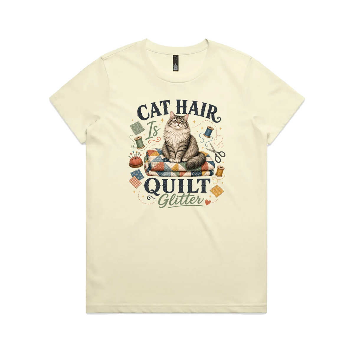 A pale butter yellow short-sleeve t-shirt with the words 'Cat Hair is Quilt Glitter' around an illustration of a cat sitting on a quilt surrounded by sewing accessories.
