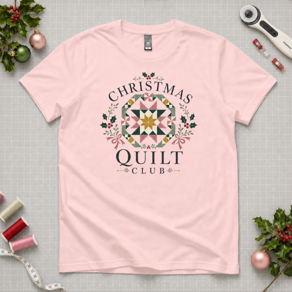 A light pink short-sleeve t-shirt with a 'Christmas Quilt Club' graphic, laid flat on a gridded cutting mat surrounded by sewing and holiday props.