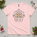 A light pink short-sleeve t-shirt with a 'Christmas Quilt Club' graphic, laid flat on a gridded cutting mat surrounded by sewing and holiday props.