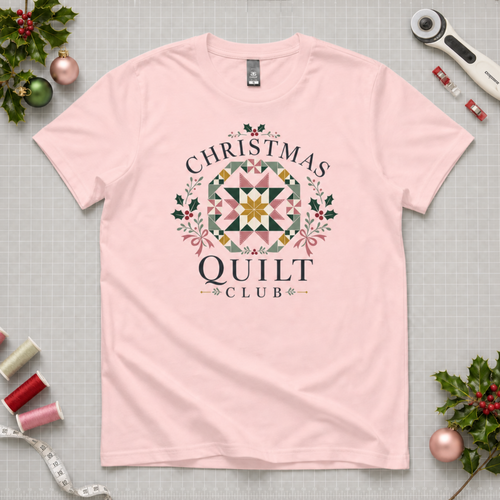 A light pink short-sleeve t-shirt with a 'Christmas Quilt Club' graphic, laid flat on a gridded cutting mat surrounded by sewing and holiday props.