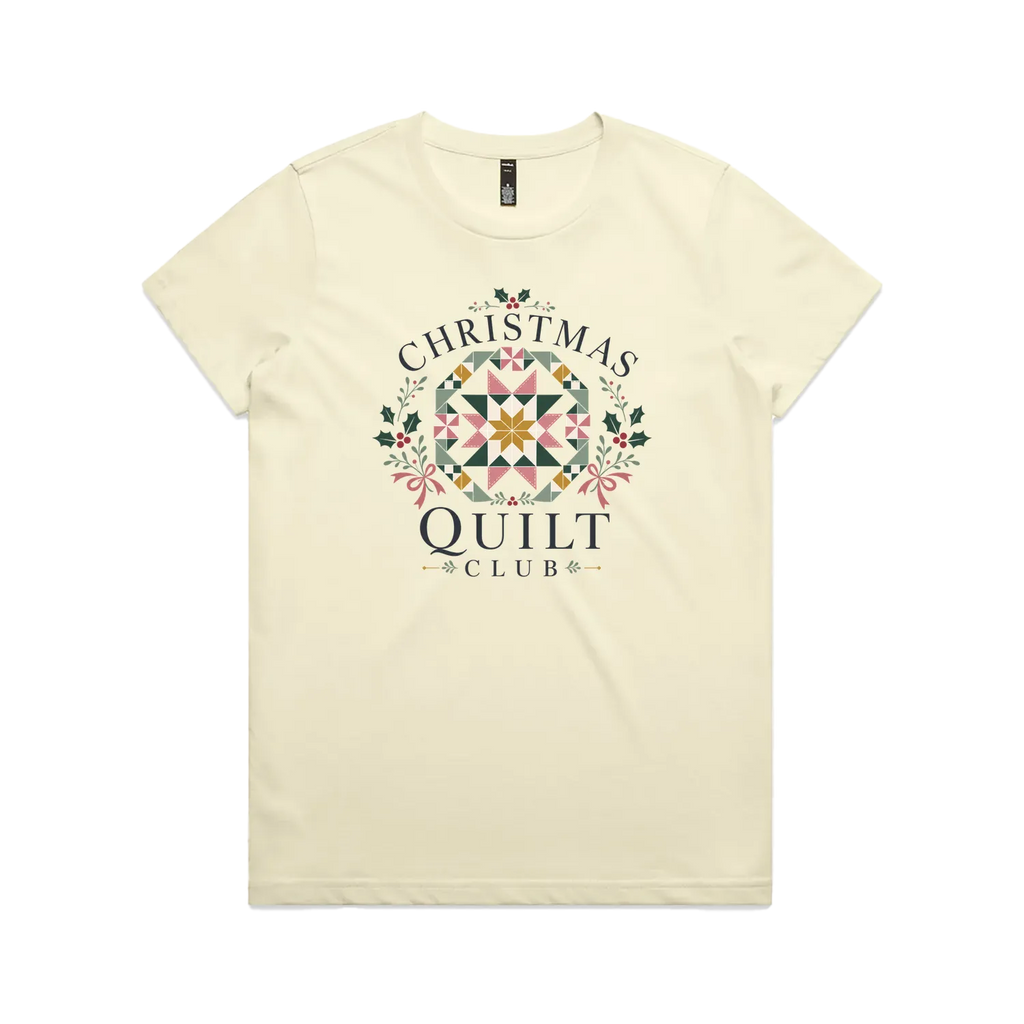A pale butter yellow short-sleeve crewneck t-shirt featuring a 'Christmas Quilt Club' graphic on the front. The graphic includes a colorful geometric quilt pattern in the center, surrounded by green holly leaves and pink bows, with the words 'CHRISTMAS QUILT CLUB' printed in dark gray.