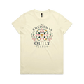 A pale butter yellow short-sleeve crewneck t-shirt featuring a 'Christmas Quilt Club' graphic on the front. The graphic includes a colorful geometric quilt pattern in the center, surrounded by green holly leaves and pink bows, with the words 'CHRISTMAS QUILT CLUB' printed in dark gray.