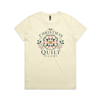 A pale butter yellow short-sleeve crewneck t-shirt featuring a 'Christmas Quilt Club' graphic on the front. The graphic includes a colorful geometric quilt pattern in the center, surrounded by green holly leaves and pink bows, with the words 'CHRISTMAS QUILT CLUB' printed in dark gray.