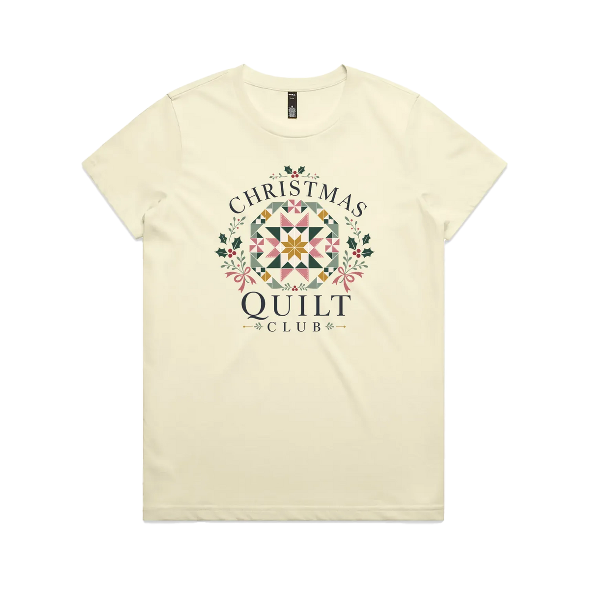 A pale butter yellow short-sleeve crewneck t-shirt featuring a 'Christmas Quilt Club' graphic on the front. The graphic includes a colorful geometric quilt pattern in the center, surrounded by green holly leaves and pink bows, with the words 'CHRISTMAS QUILT CLUB' printed in dark gray.