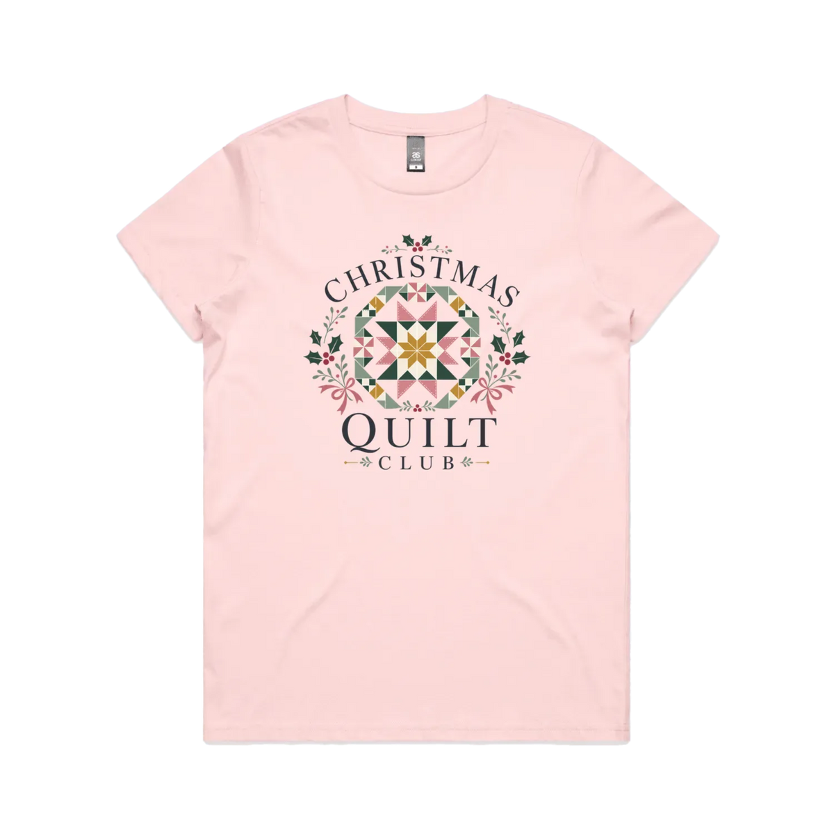 A light pink short-sleeve crewneck t-shirt featuring a 'Christmas Quilt Club' graphic on the front. The graphic includes a colorful geometric quilt pattern in the center, surrounded by green holly leaves and pink bows, with the words 'CHRISTMAS QUILT CLUB' printed in dark gray.