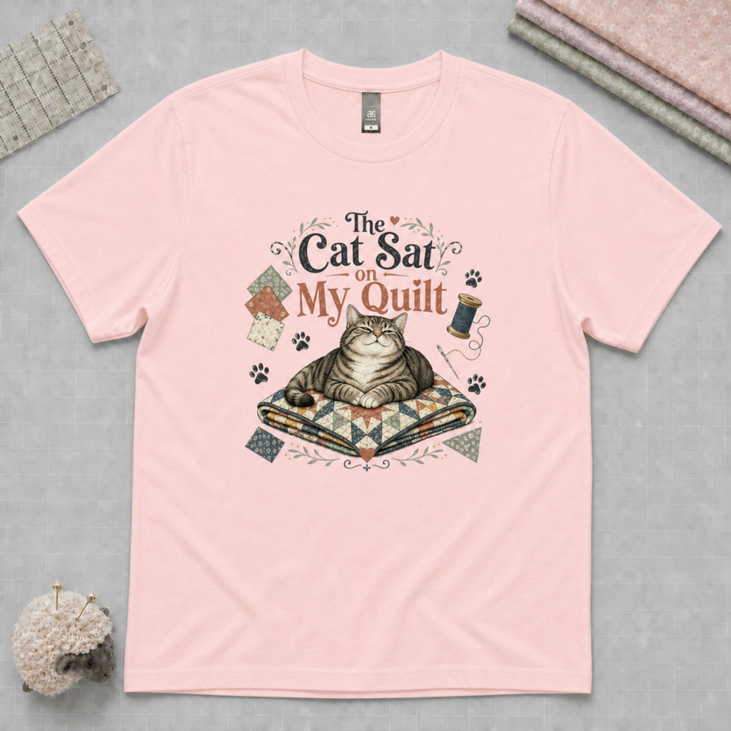 A light pink short-sleeve crewneck t-shirt with a graphic print of a cat sitting on a quilt under the text 'The Cat Sat on My Quilt'. The shirt is displayed flat on a gray cutting mat with quilting-themed props, including fabric stack and pincushion.