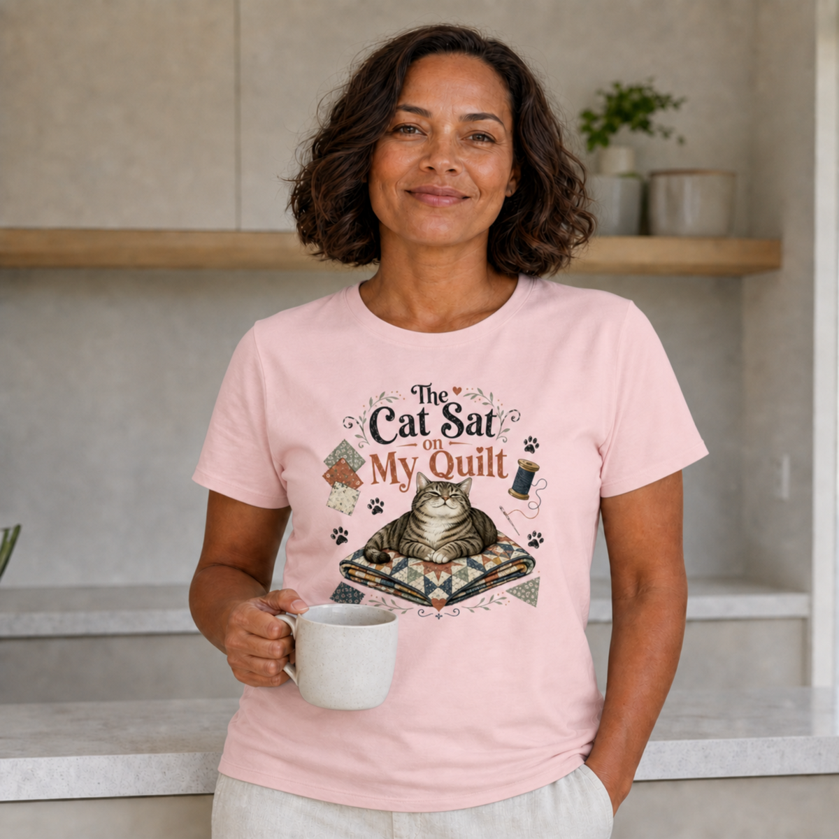 A woman wearing a light pink graphic t-shirt with a cat design and the text 'The Cat Sat On My Quilt', holding a white mug in a kitchen setting.