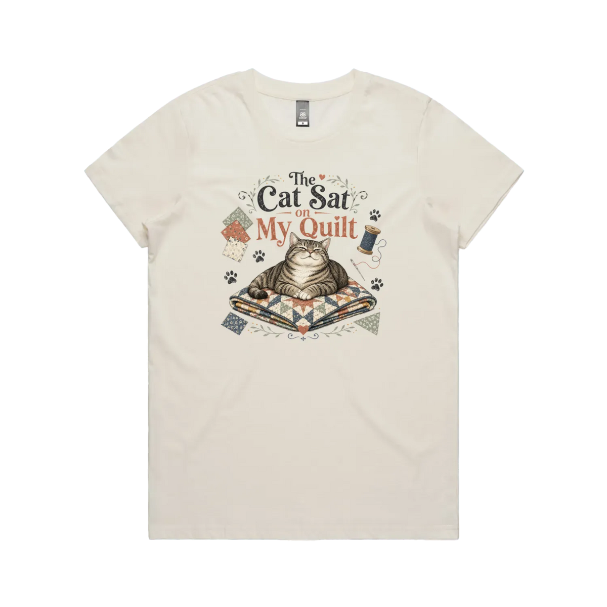 An off-white short-sleeve t-shirt with an illustration of a cat sitting on a quilt surrounded by sewing accessories and the words 'The Cat Sat on My Quilt'.