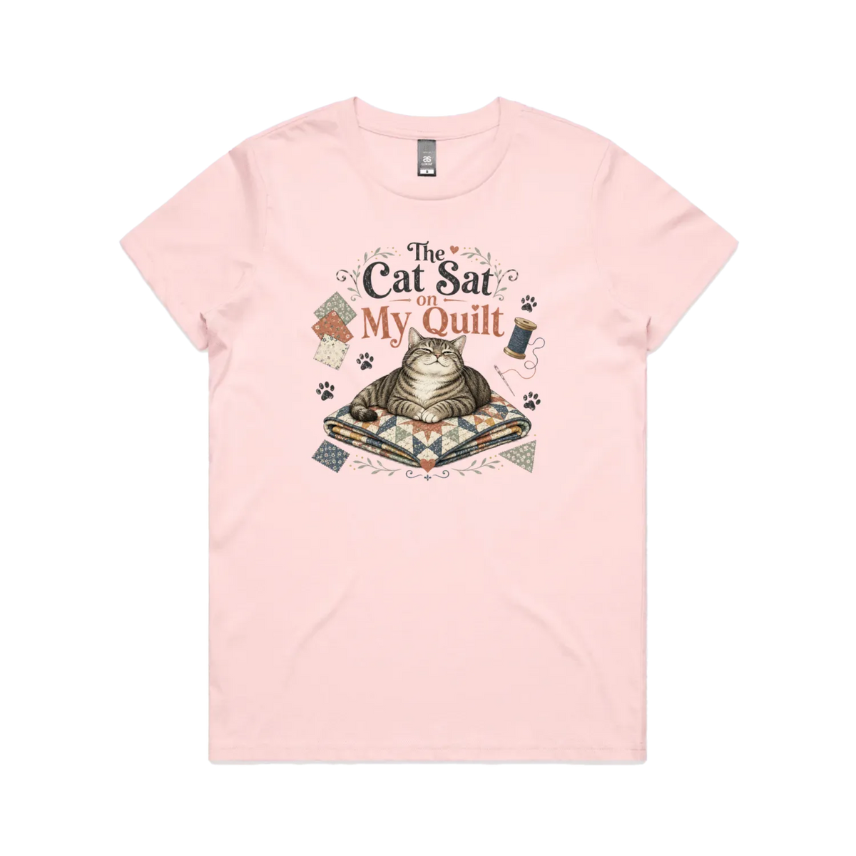 A pale pink short-sleeve t-shirt with an illustration of a cat sitting on a quilt surrounded by sewing accessories and the words 'The Cat Sat on My Quilt'.