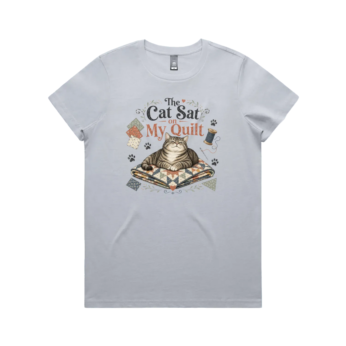 A pale powder blue short-sleeve t-shirt with an illustration of a cat sitting on a quilt surrounded by sewing accessories and the words 'The Cat Sat on My Quilt'.