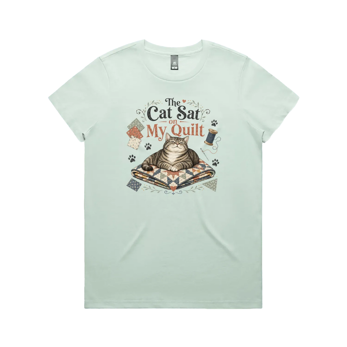A pale mint green short-sleeve t-shirt with an illustration of a cat sitting on a quilt surrounded by sewing accessories and the words 'The Cat Sat on My Quilt'.