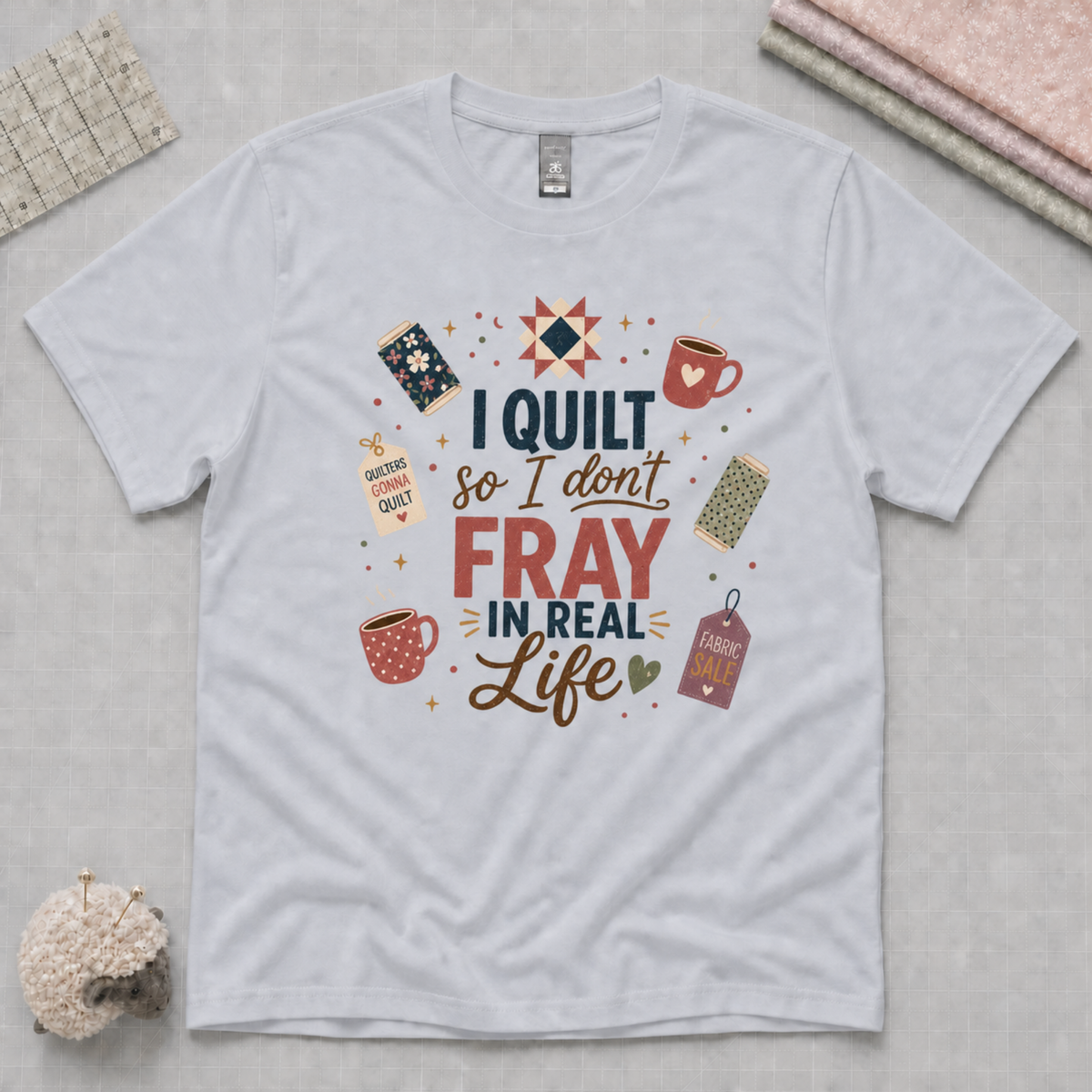 A powder blue short-sleeve t-shirt with a colorful graphic print that reads 'I Quilt So I Don't Fray in Real Life' surrounded by a quilt block, mugs, and fabric tags. The shirt is displayed flat on a light gray cutting mat with decorative props in the corners.