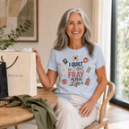 A smiling woman with gray hair sits indoors at a wooden table wearing a powder blue graphic T-shirt. The shirt has a colorful quilt-themed design with the text 'I Quilt So I Don't Fray in Real Life'.